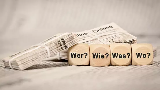 Zeitungen im Bild und Holzwürfel mit den Fragen Wer, Wie, Was, Wo