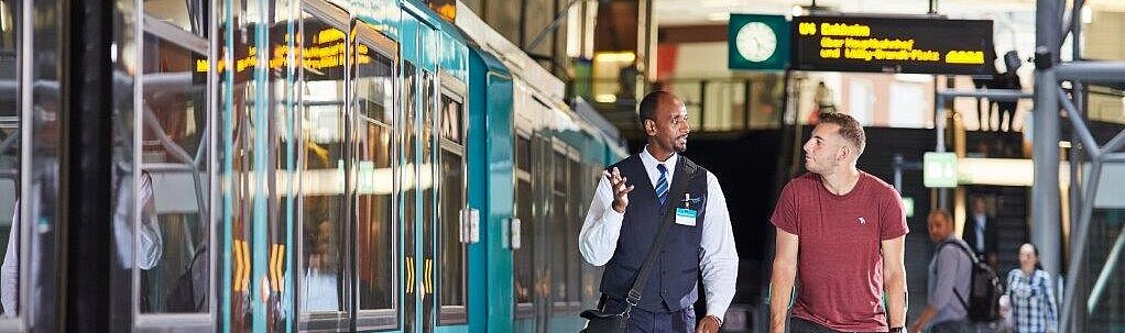 Auf der linken Seite des Bildes ist eine U-Bahn zu sehen. Rechts daneben begleitet ein Mitarbeiter des Fahrgastbegleitservice einen Kunden.