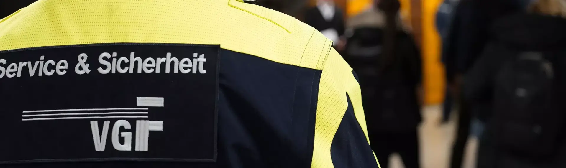 Das Bild zeigt die Rückenansicht eines Ordnungsdienstmitarbeiters in Dienstkleidung. Die Jacke trägt die Aufschrift Service und Sicherheit und das VGF Logo.