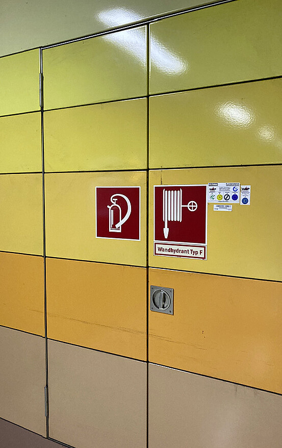Das Bild zeigt ein Piktogramm für Feuerlöscher und Hydrant an der Wand in einer U-Bahnstation.